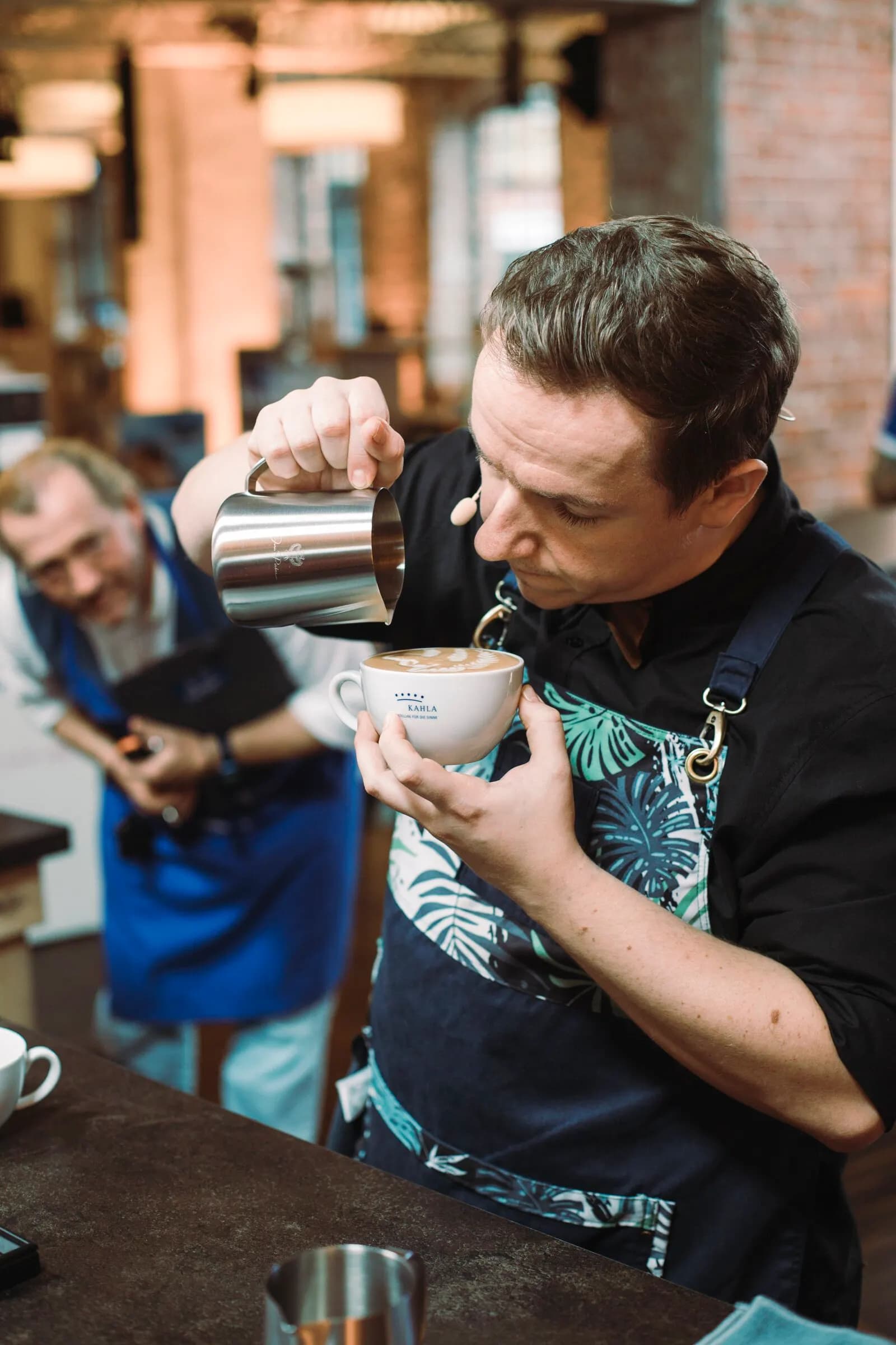 Barista gießt ein Latte-Art-Muster, im Hintergrund schaut ein Zuschauer zu